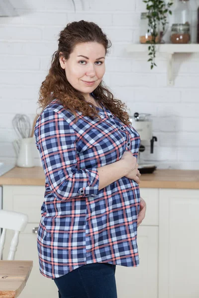 妊娠中の女性の肖像画 再生シャツ 巻き毛を着て腹を保持 キッチンインテリアに立つ — ストック写真