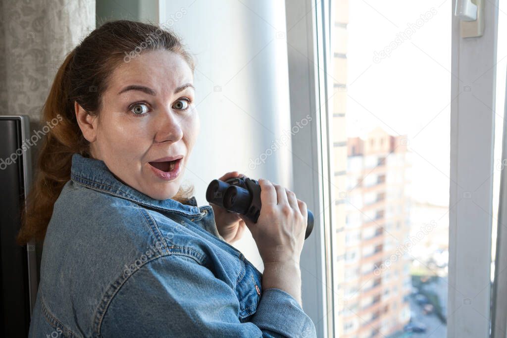 Shocked adult woman after spying out the window with binoculars, a domestic room