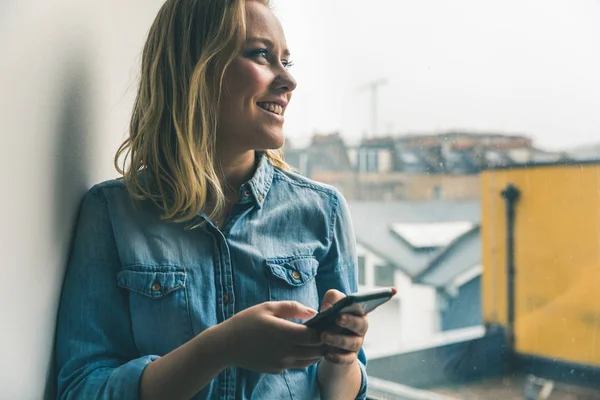 Ung kvinna med telefon tittar ut ur fönster — Stockfoto