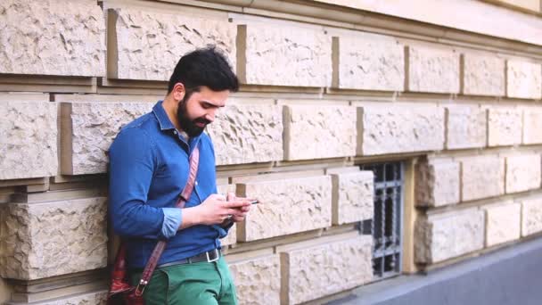 Hombre escribiendo en el teléfono inteligente y apoyado en una pared — Vídeos de Stock