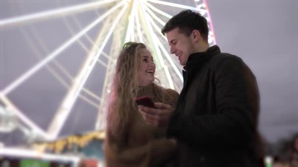 Ευτυχισμένο ζευγάρι διασκεδάζει στο λούνα παρκ και κοιτάζοντας το κινητό τηλέφωνο — Αρχείο Βίντεο