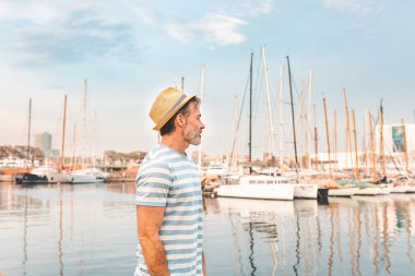 Barcelona Limanı 'ndaki insan profili portresi. Yetişkin bir adam Barcelona' da ayakta duruyor ve botlara ve gemilere bakıyor. Üzerinde deniz çizgili bir silindir ve hasır şapka var.