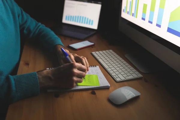 Man writing to do list at home office desk while working at night - close up of hands writing asap on a sticky note - computer and phone on the table, business and technology concepts