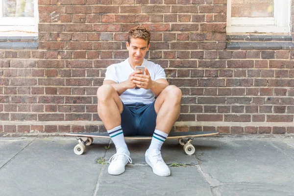 在伦敦 坐在滑板上使用电话的男人 靠墙的肖像画 一个正在放松并与社交媒体保持联系的年轻人 生活方式和技术概念 — 图库照片