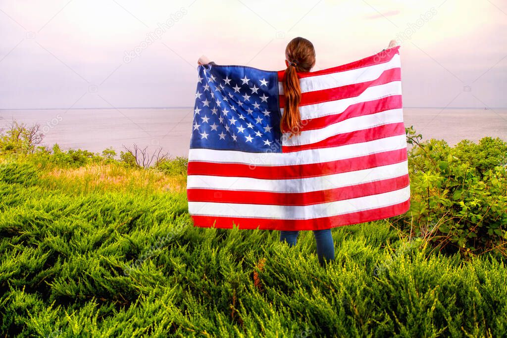 little American girl holding a star-striped US flagin her hands on the river bank 