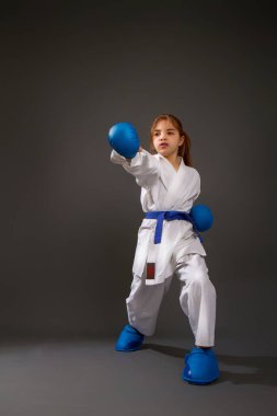 Mavi kemerli, beyaz kimonolu küçük bir kız karanlık arka planda darbeler indiriyor.