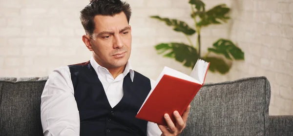 Hombre guapo está leyendo un libro mientras está sentado en un sofá en casa — Foto de Stock