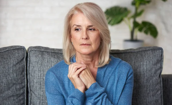 Sad senior woman is sitting on a sofa at home. old blonde lady