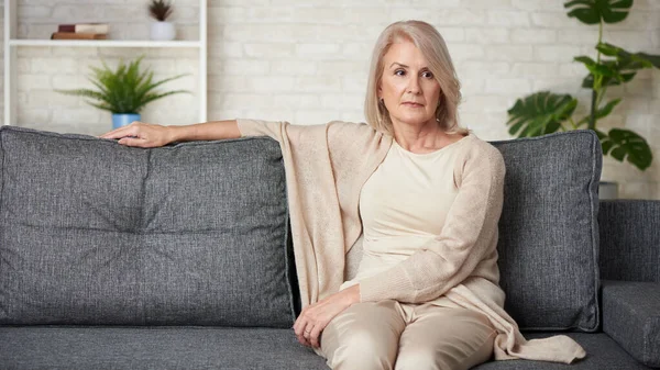 Portrait of sad older woman is sitting on a sofa at home