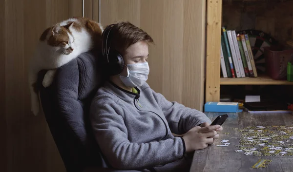 A teenager  plays a game on the phone while sitting in a chair at a table. Concept quarantine and self-isolation. Healthcare and medical concept. Pandemic virus COVID-19