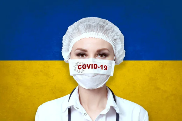戴医疗面罩的女医生 在乌克兰国旗模糊的背景上刻有Covid 19字样 大流行性病毒Covid 保健和医疗概念 — 图库照片