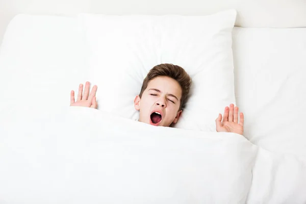 Спящий мальчик лежал в кровати, прикрываясь зевающим лицом с одеялом. — стоковое фото