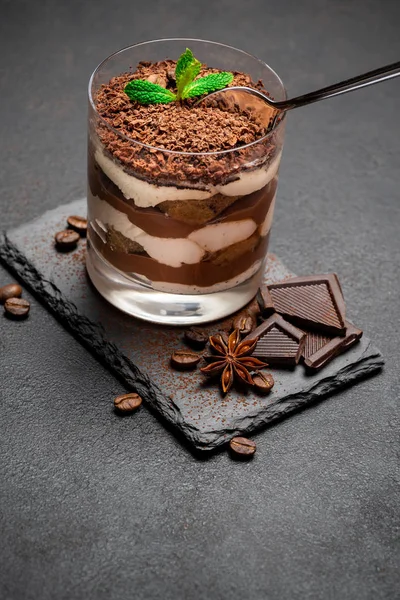 Klassisches Tiramisu-Dessert in einer Glasschale und Schokoladenstücke auf Steinschneidebrett auf darl Betonhintergrund — Stockfoto