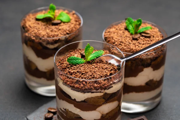 Sobremesa tiramisu clássico em uma xícara de vidro e pedaços de chocolate na placa de corte de pedra no fundo de concreto escuro — Fotografia de Stock