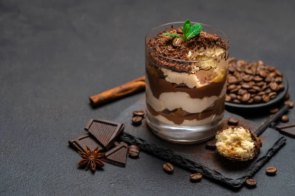 Klassisches Tiramisu-Dessert in einer Glasschale und Schokoladenstücke auf Steinschneidebrett auf dunklem Betongrund — Stockfoto