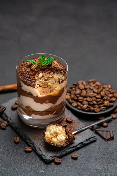 Sobremesa tiramisu clássico em uma xícara de vidro e pedaços de chocolate na placa de corte de pedra no fundo de concreto escuro — Fotografia de Stock