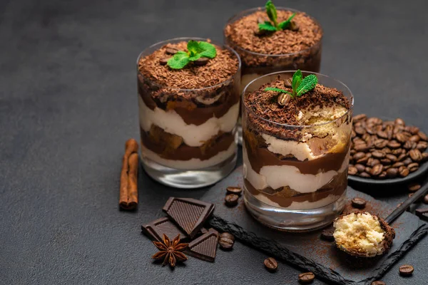 Sobremesa tiramisu clássico em uma xícara de vidro e pedaços de chocolate na placa de corte de pedra no fundo de concreto escuro — Fotografia de Stock