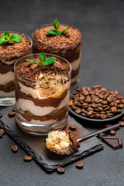 Classic tiramisu dessert in a glass cup and pieces of chocolate on stone cutting board on dark concrete background — Stock Photo, Image