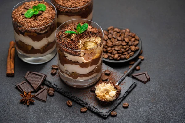 Klassisches Tiramisu-Dessert in einer Glasschale und Schokoladenstücke auf Steinschneidebrett auf dunklem Betongrund — Stockfoto