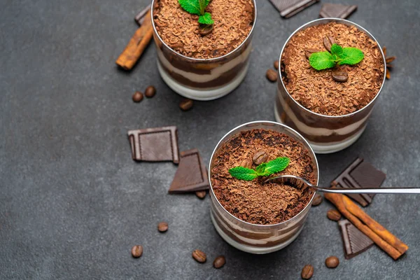 Sobremesa tiramisu clássico em uma xícara de vidro e pedaços de chocolate no fundo de concreto escuro — Fotografia de Stock