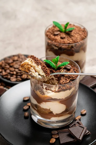 Sobremesa tiramisu clássico em uma xícara de vidro e pedaços de chocolate no fundo de concreto — Fotografia de Stock