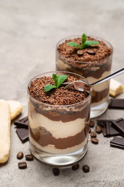 Sobremesa tiramisu clássica em uma xícara de vidro, pedaços de chocolate e biscoitos savoiardi no fundo de concreto — Fotografia de Stock