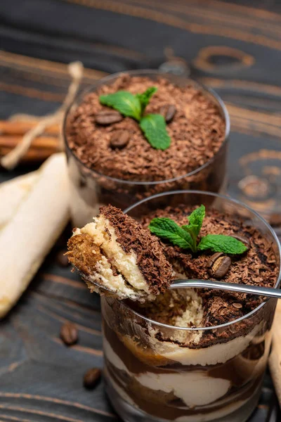 Classic tiramisu dessert in a glass cup, pieces of chocolate and savoiardi cookies on wooden background — Stockfoto