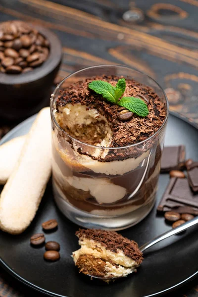 Sobremesa tiramisu clássica em uma xícara de vidro, pedaços de chocolate e biscoitos savoiardi em fundo de madeira — Fotografia de Stock