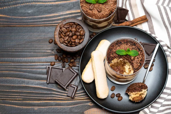 Classic tiramisu dessert in a glass cup, pieces of chocolate and savoiardi cookies on wooden background — Stockfoto