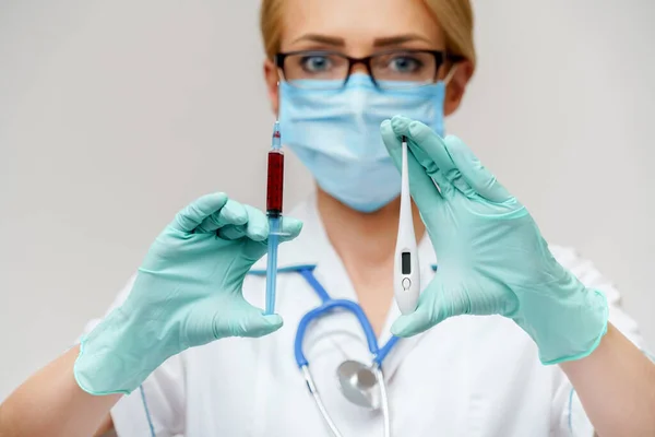 Verpleegkundige vrouw met beschermend masker en handschoenen - met elektronische thermometer en injectiespuit — Stockfoto