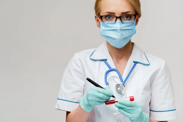 Arts verpleegkundige vrouw met beschermende masker en handschoenen - merk per pen op virus bloed reageerbuis — Stockfoto