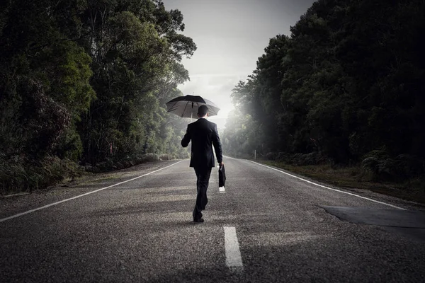 Vue arrière de l'homme d'affaires avec parapluie et valise marchant sur la route — Photo