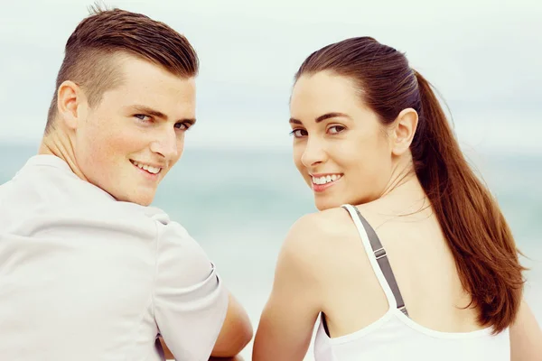 Молодая пара смотрит в камеру, сидя рядом друг с другом на пляже — стоковое фото