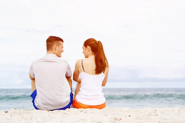 Casal jovem olhando um para o outro enquanto sentado na praia — Fotografia de Stock