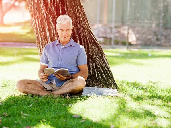 Homem sênior sentado no parque enquanto lê o livro — Fotografia de Stock