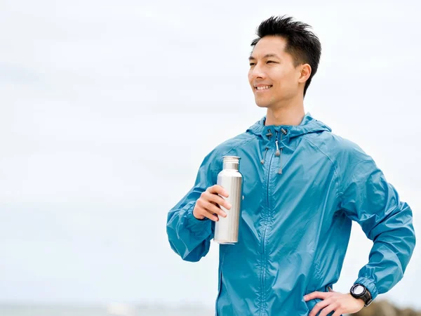 Молодой человек в спортивной одежде пьет воду после тренировки на пляже — стоковое фото