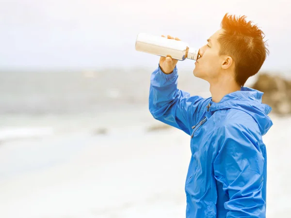 Joven con ropa deportiva bebiendo agua después del entrenamiento en la playa — Foto de Stock