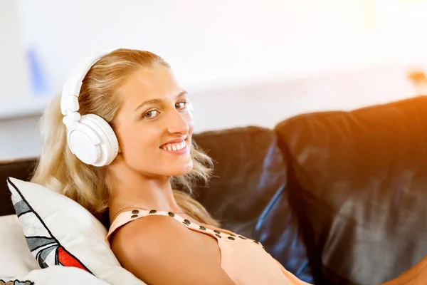 Молодая улыбающаяся женщина с наушниками отдыхает на диване, слушая музыку онлайн с помощью смартфона — стоковое фото