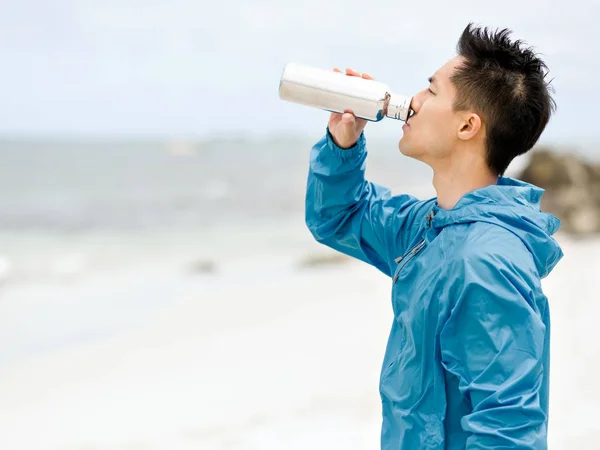 Ung man i sport kläder dricka vatten efter träningspass på stranden — Stockfoto