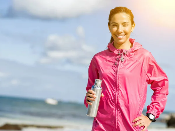 Beautiful girl in sport clothes drinking water after workout on the beach — Stock Photo, Image