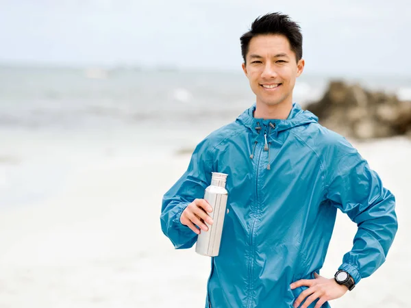 Молодой человек в спортивной одежде пьет воду после тренировки на пляже — стоковое фото