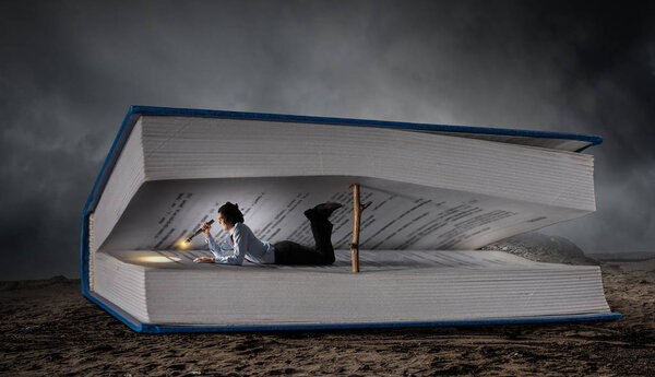 Man lying among pages of huge book with flashlight in hand. Mixed media