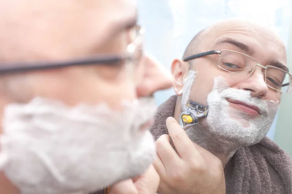 Smiling man shaving his beard at mirror in bathroom — Stock Photo, Image