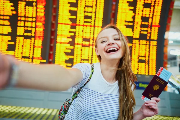 Dziewczyna w międzynarodowy port lotniczy, biorąc śmieszne selfie z paszportem i na pokład przechodzą w pobliżu tablicy informacyjnej lotu — Zdjęcie stockowe