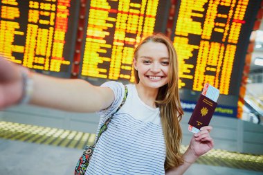 Girl in international airport, taking funny selfie with passport and boarding pass near flight information board clipart