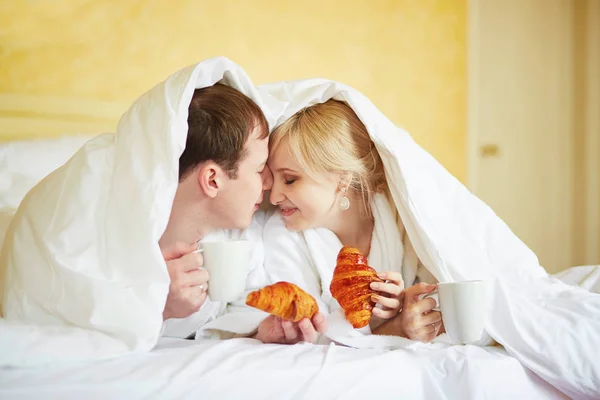 Casal em roupões brancos na cama, comendo croissants — Fotografia de Stock