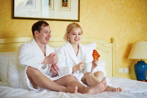 Ζευγάρι σε λευκή μπουρνούζια στο κρεβάτι, πίνοντας τον καφέ — Φωτογραφία Αρχείου