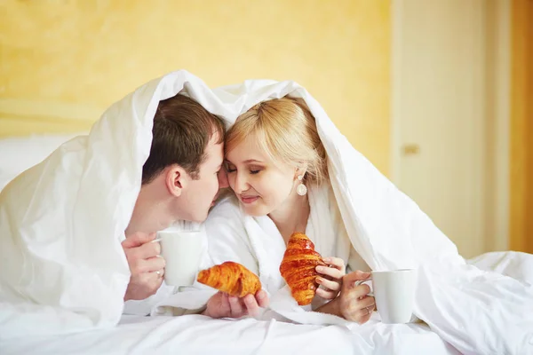 Casal em roupões brancos na cama, comendo croissants — Fotografia de Stock