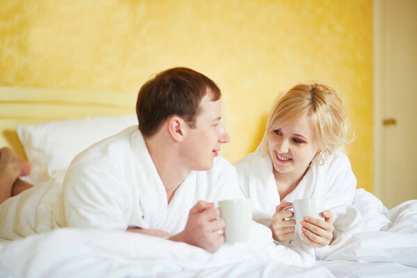 Couple in white bathrobes drinking coffee together in bed