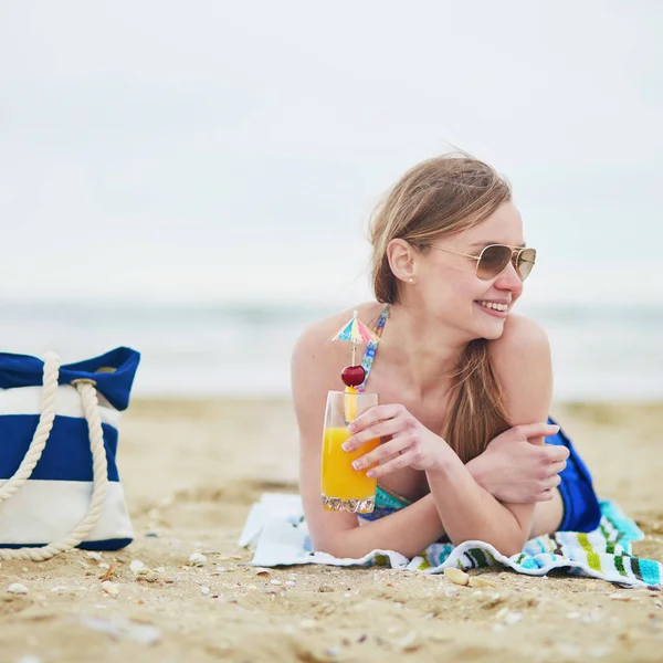 喝美味的水果或酒精鸡尾酒用纸的伞 沙滩袋沙滩附近的模型上美丽的年轻女子放松和在海滩上晒日光浴 — 图库照片